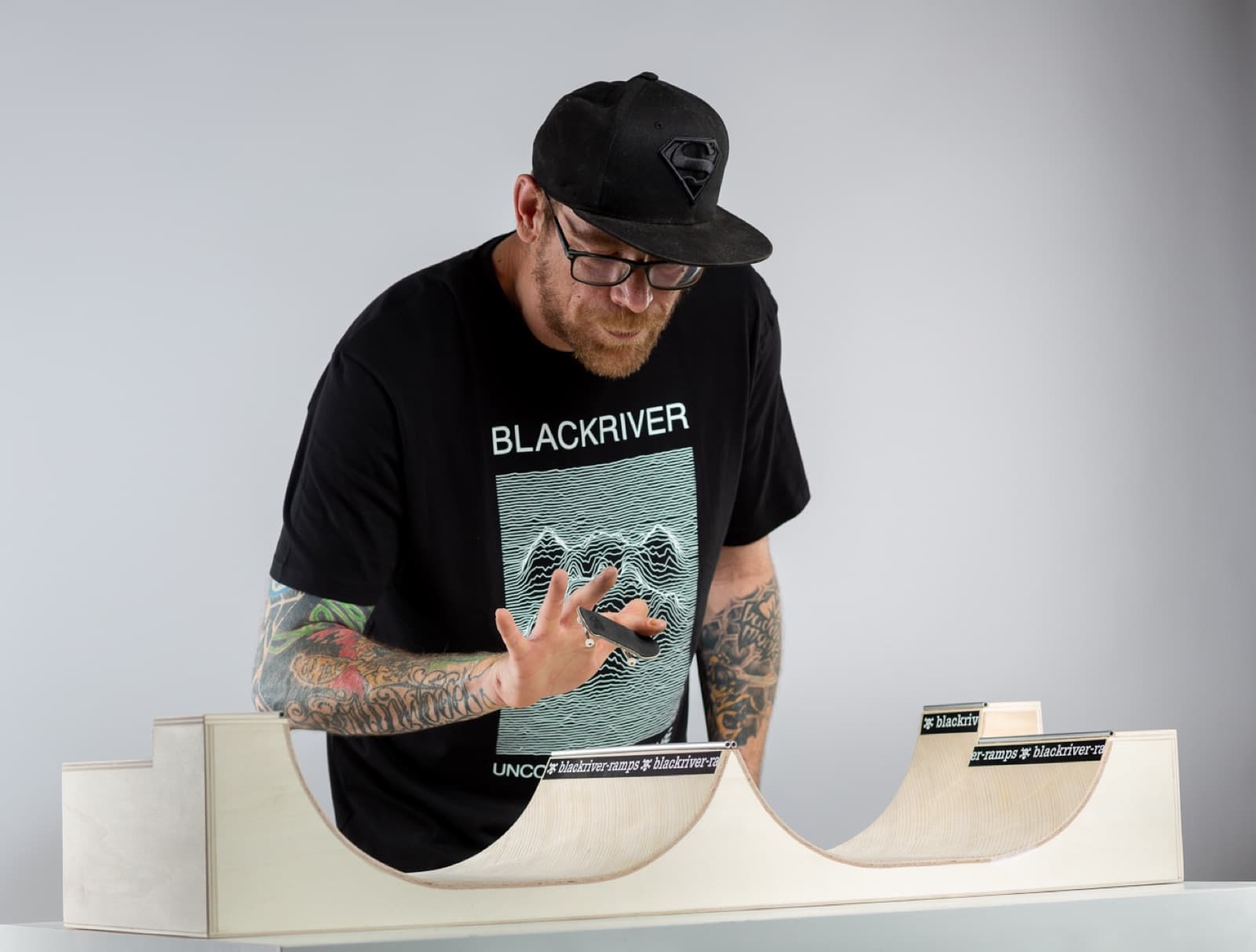 A rider flicking a fingerboard trick over a wooden Blackriver spine ramp in a studio.