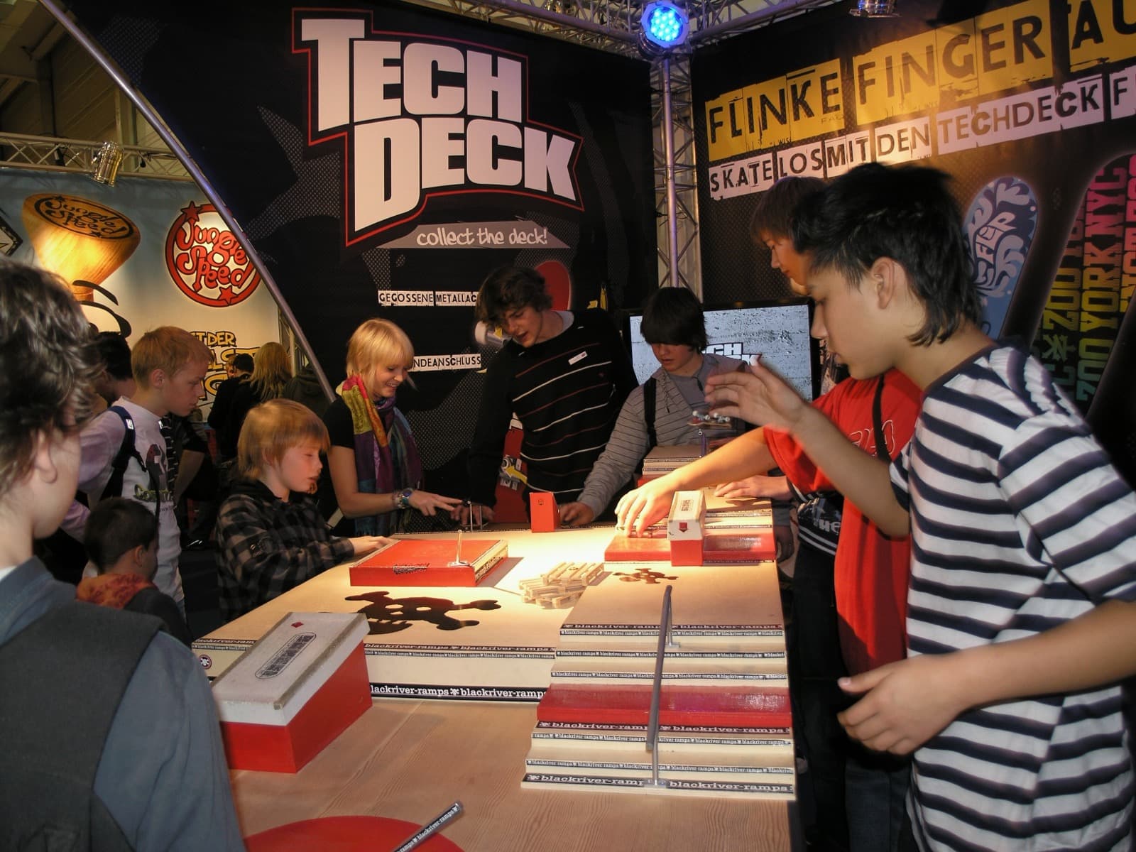 Crowded Tech Deck booth at the SPIEL fair in Essen, 2008, with kids fingerboarding on demo ramps under a huge 'collect the deck!' backdrop.