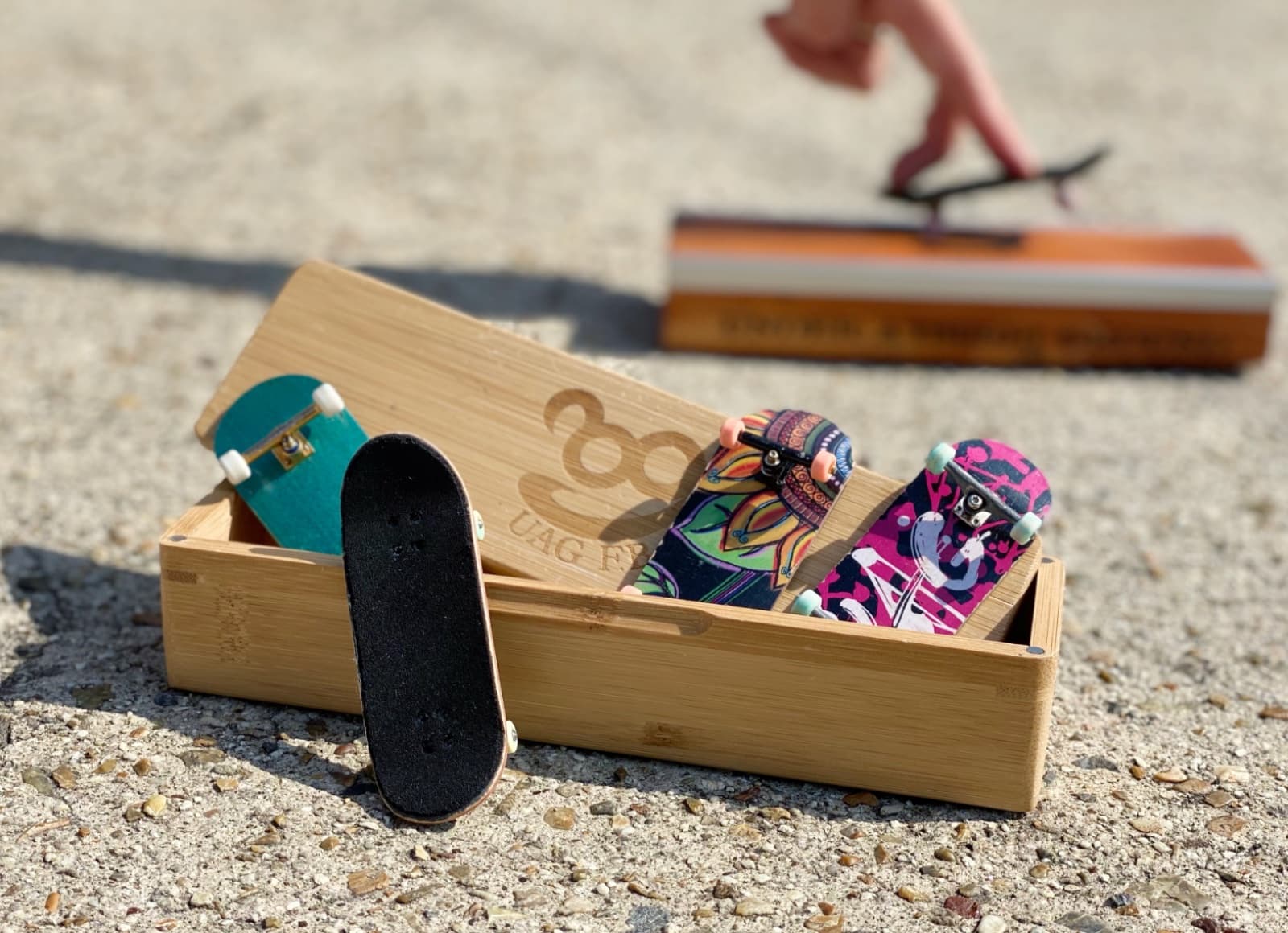 A bamboo UAG F.B deck box holding fingerboards outdoors, with a rider passing a rail in the background.