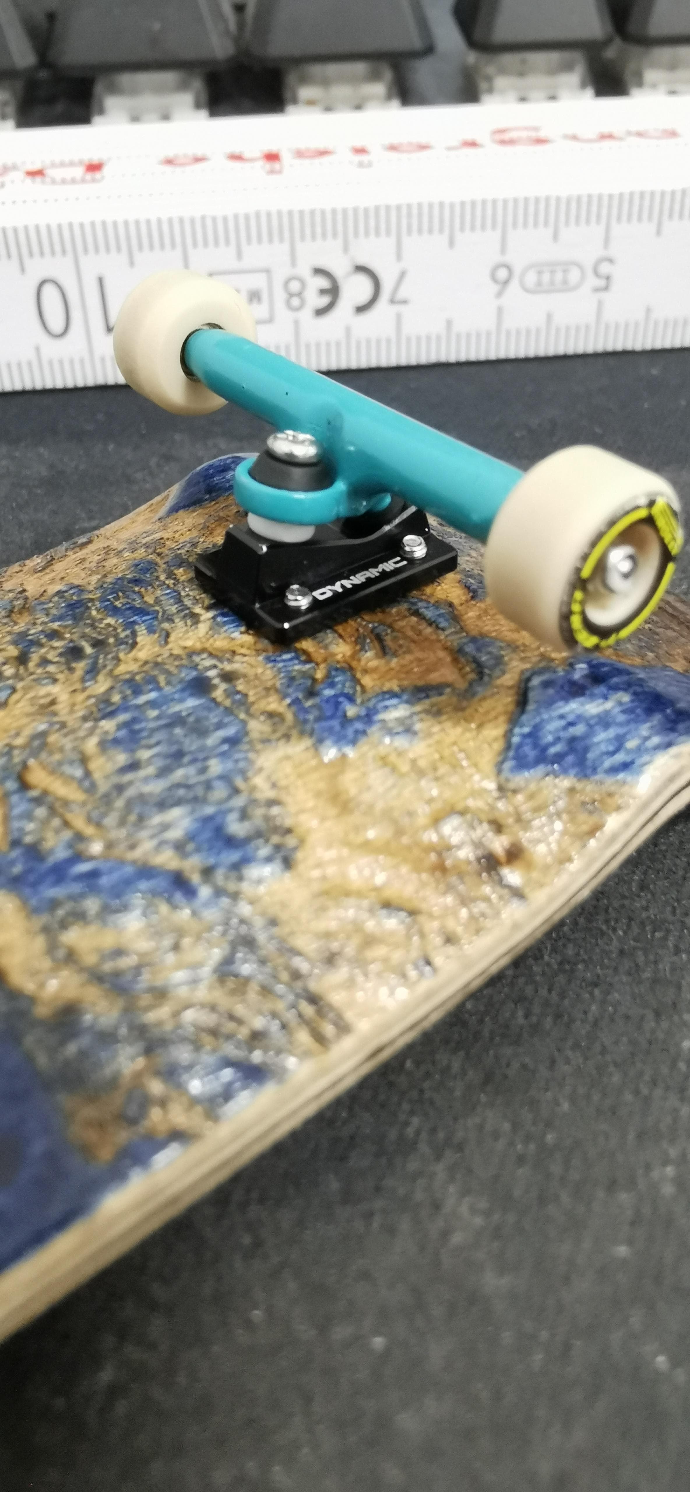 A teal fingerboard truck with cream wheels is mounted on a worn blue-and-gold deck beside a measuring ruler.